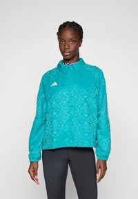 Ung kvinna med flätat hår, iklädd en tealfärgad mönstrad jacka med vitt logotyp och svarta leggings, stående mot en enkel ljus bakgrund.