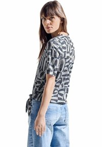Grå kortærmet T-shirt med abstrakte sort-hvide mønstre, der har en afslappet pasform og en bundet detalje i taljen. Bæres med lyseblå jeans.