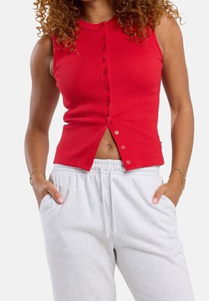 Femme portant un haut rouge sans manches à boutons partiellement déboutonné en bas et un pantalon de jogging gris clair à cordon, mains dans les poches.
