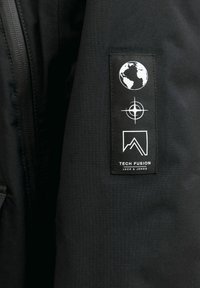 Mustmustri varrukas, millel on tikandiga patch, kus on kujutatud Maad, kompassi sümbolit, mäe kontuuri ja tekst "TECH FUSION JACK & JONES".