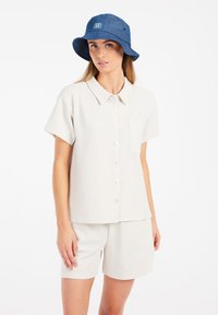Hellbeige Kurzarm-Hemd mit Knopfleiste und strukturiertem Muster, kombiniert mit passender Hose. Ergänzt mit einem blauen Bucket Hat.