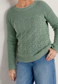 Pull en tricot vert avec un motif texturé sur le devant, manches longues et poignets côtelés, associé à un jean en denim bleu clair.