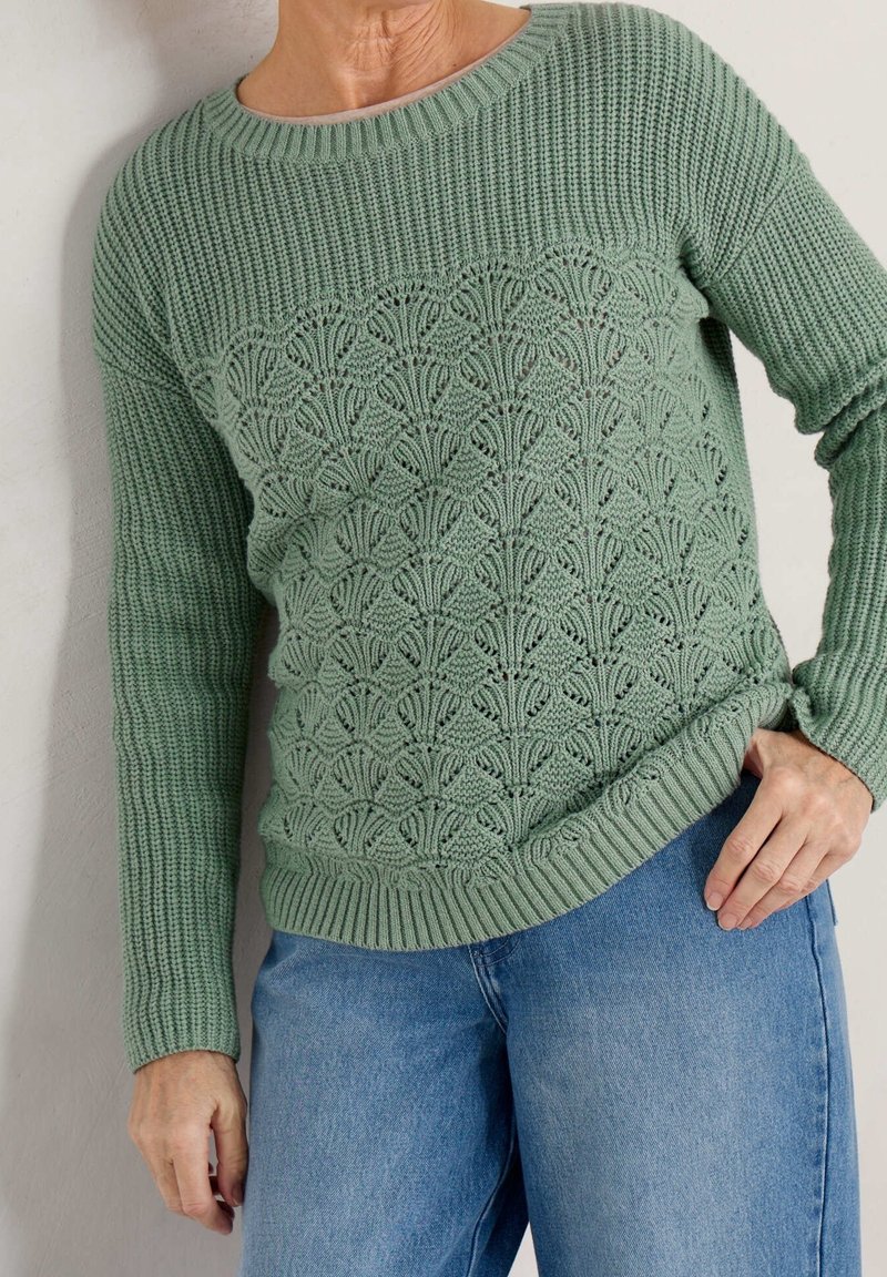 Pull en tricot vert avec un motif texturé sur le devant, manches longues et poignets côtelés, associé à un jean en denim bleu clair.