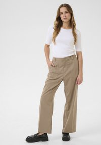 Beige vida byxor med två framfickor, i kombination med en figurnära vit ribbad topp. Modellen poserar med en hand i fickan och bär svarta loafers.