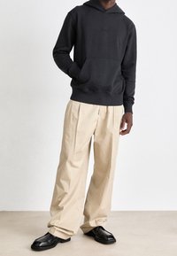 Person klädd i svart hoodie, beige vida veckade byxor och svarta fyrkantiga skor, står mot en jämn ljus bakgrund.