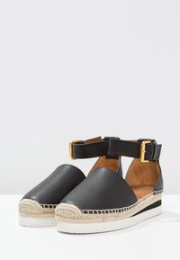 Zapatos espadrilles de cuero negro con plataforma tejida de yute, tira al tobillo con hebilla dorada y textura suave. Diseño de punta ancha y suela plana.