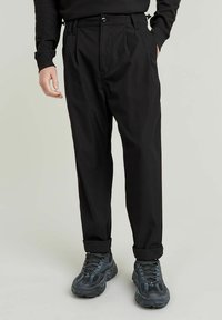 Svarta plisserade byxor med avslappnad passform, tillverkade av lätt material. Har ett knappstängning och uppvikta benslut som är kombinerade med mörka sneakers.