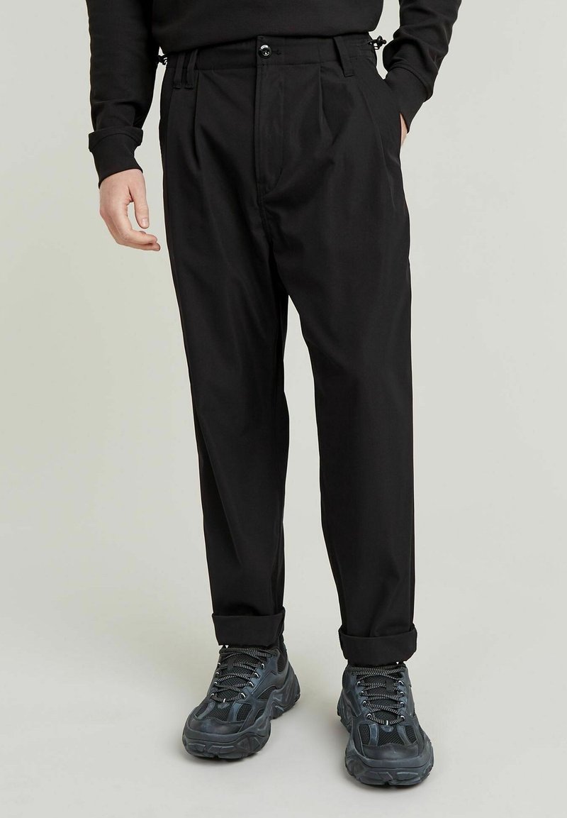 Svarta plisserade byxor med avslappnad passform, tillverkade av lätt material. Har ett knappstängning och uppvikta benslut som är kombinerade med mörka sneakers.