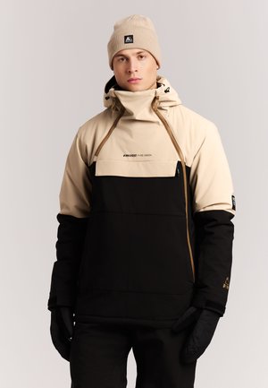 Jonge man met een beige en zwarte winterjas, een beige muts en zwarte handschoenen, staand tegen een effen lichte achtergrond.