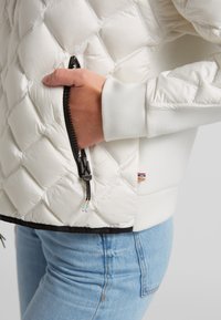 Weißer gesteppter Mantel mit glatter Textur, Reißverschlusstasche und schwarzem Reißverschlussdetail. Verfügt über einen kontrastierenden Saum und ein dekoratives Logo-Patch.