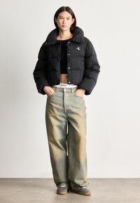 Veste en doudoune noire avec un col haut et une fermeture à pression frontale, associée à un jean en toile large bleu clair. Le modèle porte un top noir court et des chaussures grises.