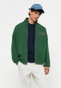 Groene zip-up jacket met hoge kraag en logo patch, gedragen over een marineblauw t-shirt. Lichtblauwe pet. Zacht fabric, relaxte fit. Witte broek.