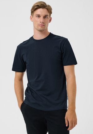 Joven con cabello castaño claro, vestido con una camiseta lisa azul marino oscuro y pantalones negros, de pie con una mano en el bolsillo contra un fondo claro.