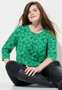 Grüne Bluse mit einem Blattmuster, runder Ausschnitt und 3/4-Ärmeln. Kombiniert mit schwarzen, enganliegenden Jeans und kniehohen schwarzen Stiefeln. Glatte Textur.