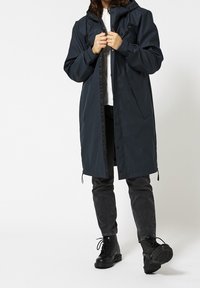 Marinblå vattentät långrock med huva, försedd med dragkedja och tryckknappsstängning, justerbara dragskor och sidofickor. Kombinerad med svarta jeans och stövlar.