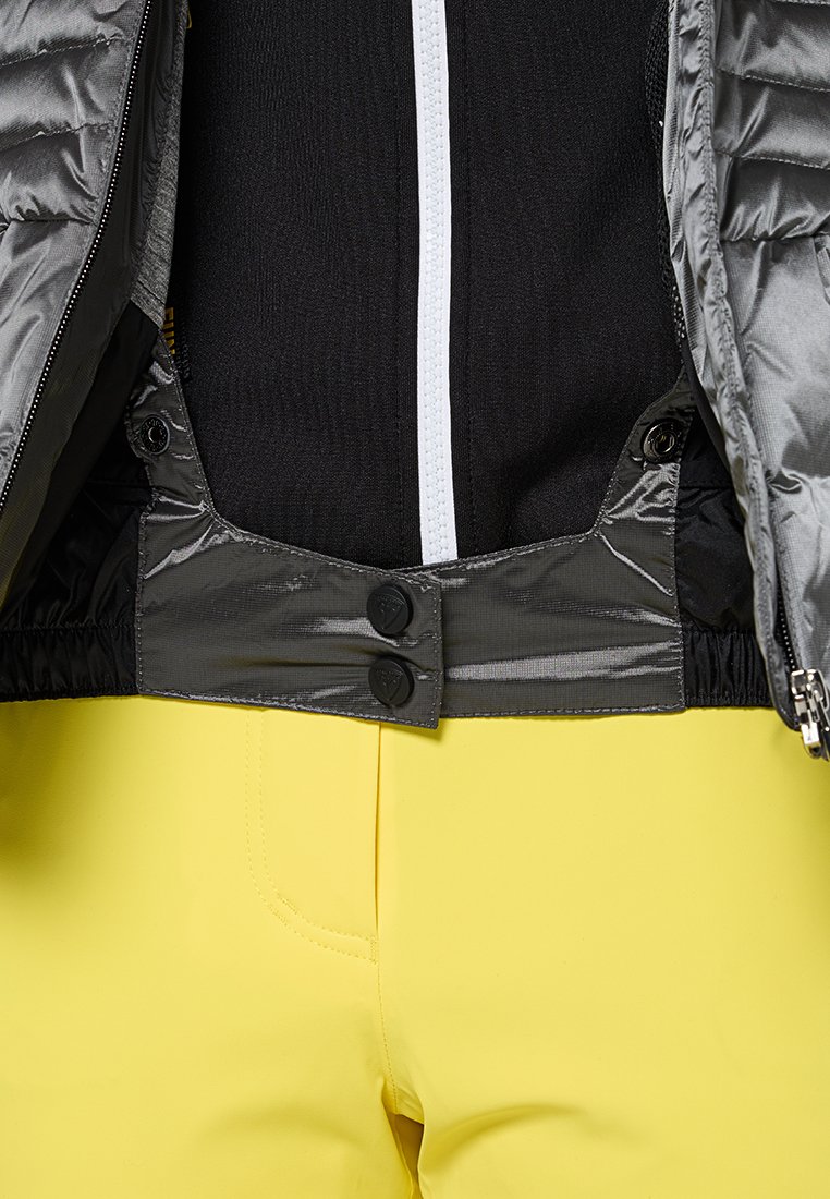 Chaquetón gris acolchado con cremallera y dos botones negros en la cintura, combinado con pantalones amarillos brillantes. Tejido texturizado y diseño ajustado.