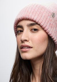 Beanie rosa lavorato a maglia con una texture a coste e una banda bianca. Presenta un'etichetta piccola di lato. Visione ravvicinata, mostrando i dettagli del viso.