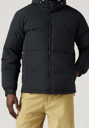 Homme portant une doudoune noire à capuche zippée, les mains dans les poches, associée à un pantalon beige sur un fond uni.