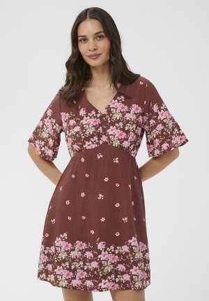 Žena oblečená v hnedých krátkych rukávoch s ružovými a bielymi kvetinovými vzormi, stojaca pred jednoduchým svetlým pozadím.