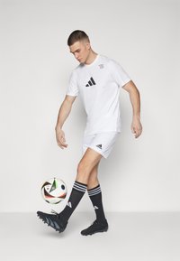 Maglietta atletica bianca con logo nero, pantaloni corti bianchi, calze da calcio nere con strisce bianche, scarpe da calcio nere e un pallone da calcio colorato.