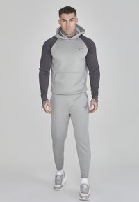 Uomo con tatuaggi che indossa un hoodie grigio con maniche nere, pantaloni felpati grigi abbinati e scarpe grigie e bianche, in piedi contro uno sfondo neutro.