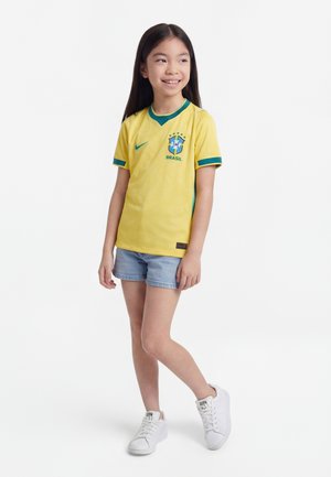 Mlada deklica stoji in se smehlja, oblečena v rumeno nogometno majico Brazilije, jeans kratke hlače in bele superge na enobarvni beli podlagi.