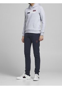 Šedá mikina s kapucí a logem, přední kapsou a žebrovanými manžetami. Námořnické tepláky s vyšívanými detaily, zúžený střih. Bílé tenisky s barevnými pruhy.