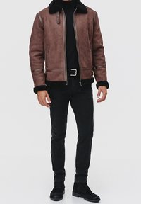 Giacca in suede marrone con colletto in pelliccia sintetica, zip frontale e accenti grigi. Indossata sopra un dolcevita nero, abbinata a pantaloni slim-fit neri e scarpe.