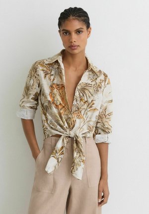 Femme aux cheveux tressés portant un pantalon beige taille haute et une chemise safari manches longues nouée avec un imprimé lion et feuillage, debout contre un mur blanc.
