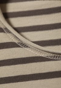 Beige stof met donkere horizontale strepen; heeft een ronde halslijn en eenvoudige stiksels langs de randen. Zachte textuur.