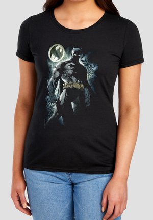 Frau trägt ein schwarzes T-Shirt mit Batman-Grafik und Fledermaussignal im Hintergrund, kombiniert mit hellblauen Jeans.