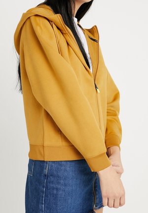 Person iført en sennepsgul hættetrøje med lynlås og hætte og en blå denim nederdel, hænderne foldet foran.