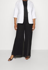 Bílý blazer s tříčtvrtečními rukávy přes černý top, doplněný černými širokými kalhotami s transparentními bočními panely. Černé sandály.
