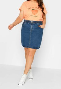 Denimová sukně se zvýrazněným tvarem, elastickým pasem a zapínáním na knoflík. V kombinaci s růžovým tričkem s grafikou a bílými vysokými teniskami.