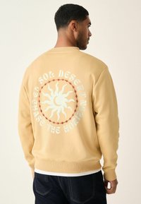 Beige sweatshirt med lange ærmer, ribbede manchetter og et stort solgrafik på ryggen. Teksten lyder "JAGT HORIZONTEN" i kontrasterende farver.
