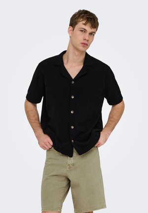 Jeune homme aux cheveux courts portant une chemise noire à manches courtes boutonnée et un short beige jusqu'aux genoux, mains dans les poches, fond neutre.