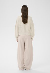 Frau mit langem braunem Haar, die einen cremefarbenen Strickpullover und weite beige Hosen trägt, und mit dem Rücken zu einer weißen Wand steht.