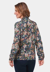 Blusa floreale a maniche lunghe di colore blu scuro con intricati motivi colorati, colletto arricciato e polsini con bottoni. Tessuto leggero con una consistenza morbida.