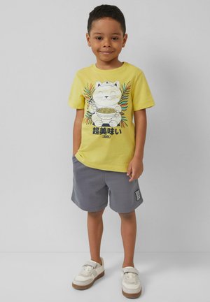 Żółta bawełniana koszulka z grafiką kota trzymającego makaron, w połączeniu z szarymi szortami. Sneakersy mają białą cholewkę i brązową podeszwę.
