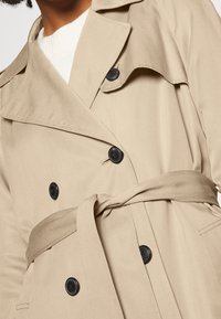 Vaalean beige-trenssi takki, jossa on tuplapainike etuosa, mustat nappit ja sidottava vyö. Ominaisuuksiin kuuluu klassinen kaulus ja sileä pinta.