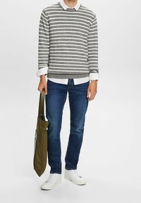 Pull gris et blanc à rayures sur une chemise blanche, jean bleu et baskets blanches. Tient un sac fourre-tout vert olive. Design simple.