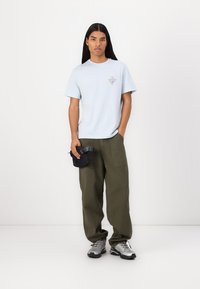 T-shirt en coton bleu clair avec un graphique en triangle, associé à un pantalon ample vert olive et des baskets argentées. Un petit pouch noir est tenu à la main.