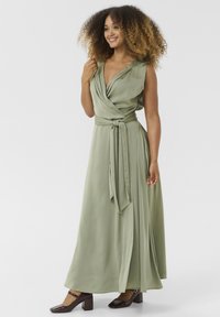 Robe enveloppante verte sans manches avec un décolleté en V profond, taille cintrée et silhouette fluide. Le mannequin porte des chaussures à talon bloc en cuir laqué noir.