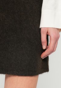 Χέρι resting κατά κόντρα textured φούστα, λευκή μπλούζα με μακριά μανίκια ορατή στον καρπό, τόνος δέρματος ανοιχτός, νύχια κοντά και φυσικά.