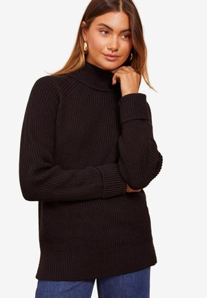 Mørkebrun ribbet strik turtleneck sweater med lange ærmer, der har en afslappet pasform og tykke manchetter, parret med blå jeans.