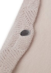 Lichtbeige stof met een gebreide textuur, met een metalen knoop en een gladde achtergrondstof. Het breipatroon is zichtbaar langs de rand.