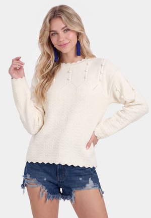 Lyshåret kvinde iført en cremefarvet tekstureret sweater med pomponer og blå kvast øreringe, kombineret med slidte denimshorts.