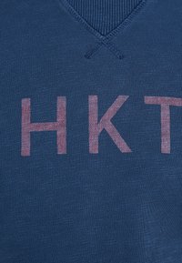 Gros plan d’un tissu bleu avec des lettres roses "HKT" imprimées, montrant un détail d'encolure en V côtelée et un motif tricoté texturé.