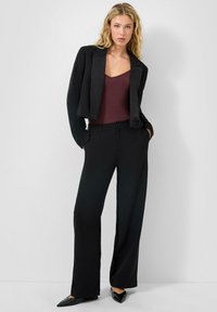 Blazer noir court sur un débardeur bordeaux, associé à un pantalon large noir. Un tissu texturé et des ballerines noires pointues complètent le look.