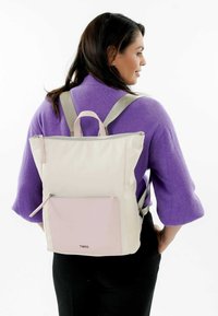 Zaino beige con una tasca frontale rosa, chiusura a zip e spallacci grigi. Presenta una maniglia superiore e un branding minimale sul fronte.